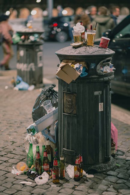 A black public waste bin situated on a cobbled sidewalk, visibly overflowing with various refuse. The lid is open and topped with discarded fast-food cups, paper food containers, and a plastic cup, some with straws and lids still attached, indicating recent use. Around the base of the bin, there are numerous empty glass bottles, primarily green and brown, scattered among crumpled paper, tissue, and additional plastic waste. A black trash bag leans against the side of the bin, partially filled with rubbish, and a cardboard box is wedged into the side opening of the bin, overflowing with waste. In the background, blurred scenes of an urban environment are visible, including parked cars and pedestrians, with trees and buildings further beyond. The scene is illuminated by natural daylight, with a somewhat cluttered and untidy appearance, reflecting a common sight in public spaces that may involve casual or immediate disposal outside designated waste collection points. Rubbish Clearance Ealing often handles the removal of such street litter and waste accumulation, supporting responsible disposal and cleaner private or public areas.