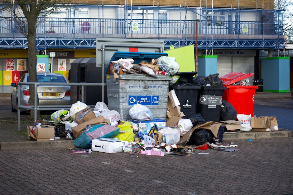 An outdoor scene showing an overflowing collection of mixed rubbish and waste materials gathered around and on top of two large outdoor recycling containers, one grey and one black, with a bright red waste bin nearby. The rubbish includes cardboard boxes, paper, plastic bags, packaging materials, and miscellaneous household waste, some spilling onto the paved ground and sidewalk. The containers are situated on a paved area next to a metal railing, with a parked vehicle partially visible on the left side. In the background, there is a building under construction or renovation, covered in blue scaffolding and mesh, and storefronts at street level displaying signs and logos. The lighting indicates daytime with natural light, highlighting the cluttered and disordered state of the waste collection, illustrating a typical scenario of unmanaged rubbish accumulation that could be managed through services such as those offered by Rubbish Clearance Ealing, supporting alternative waste disposal methods outside local authority collection.