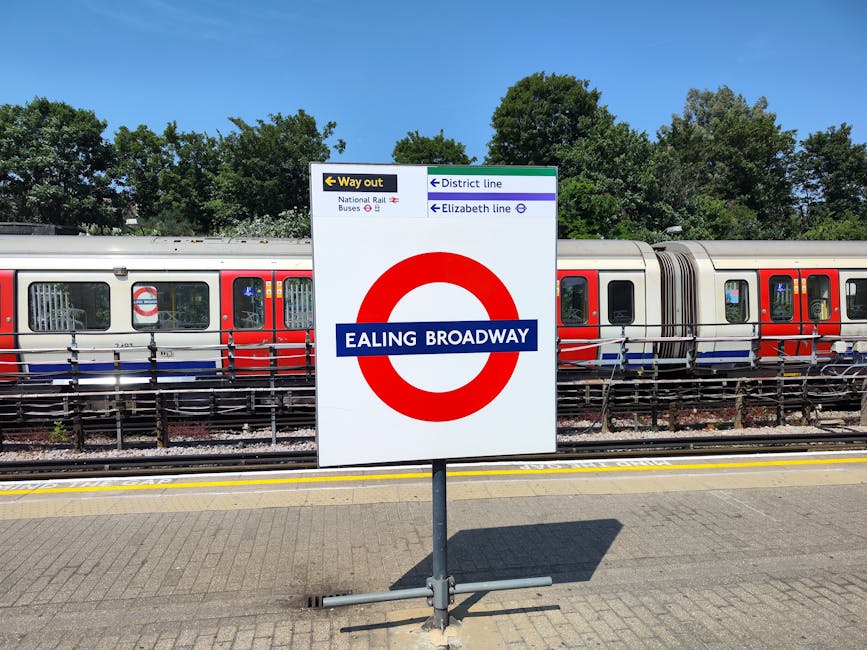 A white rectangular sign with a red circle and blue horizontal stripe displaying the text 'EALING BROADWAY' in white letters is positioned on a metal pole at a train station platform. Behind the sign, a modern London train with a metallic silver body and red and blue stripes runs along the railway tracks, partially obscured by a black metal safety railing. The train has multiple carriages with numerous rectangular windows, some reflecting the sky and surrounding trees. The platform surface is paved with light grey bricks, and a shadow cast by the sign falls onto the ground. In the background, behind the train, there is a row of lush green trees under a clear blue sky, suggesting a bright, sunny day. The scene captures a typical urban environment where public transportation infrastructure is visible, with the signage indicating station information and directions relevant to commuters and visitors. Occasionally, such signs are seen near delivery or collection points for waste management services when moving luggage or managing station waste, though in this specific context, it strictly shows a typical train station sign.