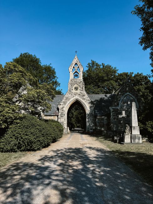 The image depicts a historic stone church or chapel with a tall, ornate steeple topped with a cross, situated within a green landscape under a clear blue sky. The building is constructed from grey stone with a pointed arch entrance and a circular clock face on the steeple. Flanking the entrance are mature, leafy trees, casting shadows on the gravel pathway leading up to the church. To the right of the arched gateway, there are stone monuments or memorial plaques mounted on the wall, suggesting a heritage or burial site. The scene appears peaceful, with natural sunlight illuminating the weathered textures of the stonework and lush foliage. While the focus is on the historic religious structure, the surrounding environment indicates it might be part of a historic churchyard or garden. Recognising the need for alternative or private waste management options, this setting naturally aligns with the idea of on-site clearance or independent rubbish disposal, consistent with services provided by Rubbish Clearance Ealing in the local area, although no waste materials are visible in this specific image.