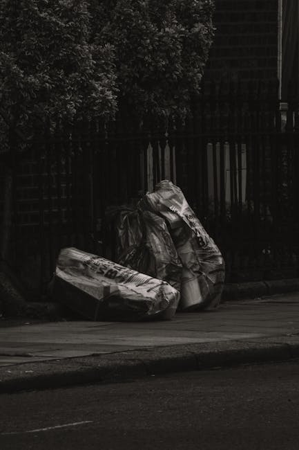 Three large black trash bags filled with rubbish are placed on the pavement near a black metal fence, with dense foliage visible behind the fence. The bags appear to be made of shiny, durable plastic and are tightly packed, with some showing creases and folds. They are positioned close together, leaning slightly to one side, and are situated outside what appears to be a residential or urban area. The scene is captured in natural daylight, casting soft shadows on the ground. This setup suggests a private rubbish collection or on-site clearance, possibly handled by services such as Rubbish Clearance Ealing, and highlights the importance of proper disposal methods for household waste. The surrounding environment is relatively quiet, with a curb marking the edge of the street, emphasizing that the waste is temporarily stored for collection or removal as part of general rubbish management in the area.
