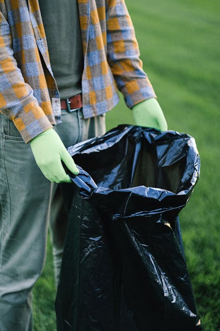 A person wearing a plaid shirt, grey trousers, and bright green gloves is holding open a large black plastic rubbish bag outdoors on a grassy area. The bag appears to be empty or lightly filled, with its top open and slightly crumpled. The background features a blurred green lawn, suggesting the scene takes place in a garden or park setting. The person’s posture indicates they are in the process of discarding waste, aligned with waste collection or rubbish removal activities. This scene exemplifies private waste handling or on-site clearance, potentially performed by a rubbish removal service such as Rubbish Clearance Ealing, to manage waste in a controlled and environmentally responsible manner, distinct from local authority rubbish collection services.