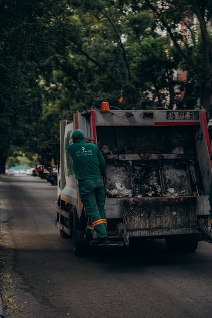 A worker dressed in green uniform and safety helmet is standing at the rear of a white and red rubbish collection vehicle on a suburban street during daytime. The worker is actively engaged with the open rear compartment of the truck, which appears to be used for waste collection and has visible dirt, stains, and accumulated debris inside. The vehicle is parked on the curb alongside a tree-lined pavement, with green foliage overhead indicating a residential or mixed-use area. In the background, other vehicles are visible parked along the street, with some greenery and parts of buildings faintly seen through the trees. Natural daylight illuminates the scene, highlighting the textures of the truck's metal surfaces and the pavement, which is slightly worn. This scene exemplifies an independent waste collection operation, reflecting private rubbish removal services that offer same-day collection options, as managed by Rubbish Clearance Ealing.
