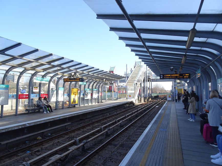 A modern railway station platform with a curved, transparent roof structure supported by metal beams extends along the length of the platform, providing shelter for waiting passengers. The platform surface is made of concrete with tactile paving strips near the edge for safety. On the left side, there are a few seated passengers against a wall with informational posters and electronic information displays showing train schedules; some are engaged in conversation, while others are focused on their phones. To the right, several individuals stand or sit with luggage, including a person with a pink rolling suitcase, waiting for a train. In the background, a staircase leads up to an overpass, and behind the platform, there are urban buildings and a clear, blue sky indicating daytime. The scene is well-lit with natural sunlight, creating a clear and calm atmosphere, typical of a public transportation hub supporting private or independent travel, such as rubbish removal or waste disposal needs addressed through local services like Rubbish Clearance Ealing.
