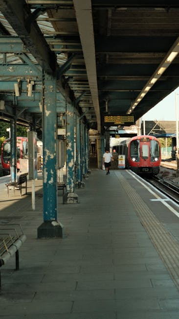 A modern railway station platform with a curved, transparent roof structure supported by metal beams extends along the length of the platform, providing shelter for waiting passengers. The platform surface is made of concrete with tactile paving strips near the edge for safety. On the left side, there are a few seated passengers against a wall with informational posters and electronic information displays showing train schedules; some are engaged in conversation, while others are focused on their phones. To the right, several individuals stand or sit with luggage, including a person with a pink rolling suitcase, waiting for a train. In the background, a staircase leads up to an overpass, and behind the platform, there are urban buildings and a clear, blue sky indicating daytime. The scene is well-lit with natural sunlight, creating a clear and calm atmosphere, typical of a public transportation hub supporting private or independent travel, such as rubbish removal or waste disposal needs addressed through local services like Rubbish Clearance Ealing.